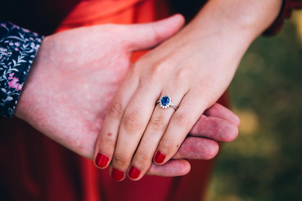 A man’s hand holding his bride-to-be’s, her hand adorned with a sapphire halo engagement ring.