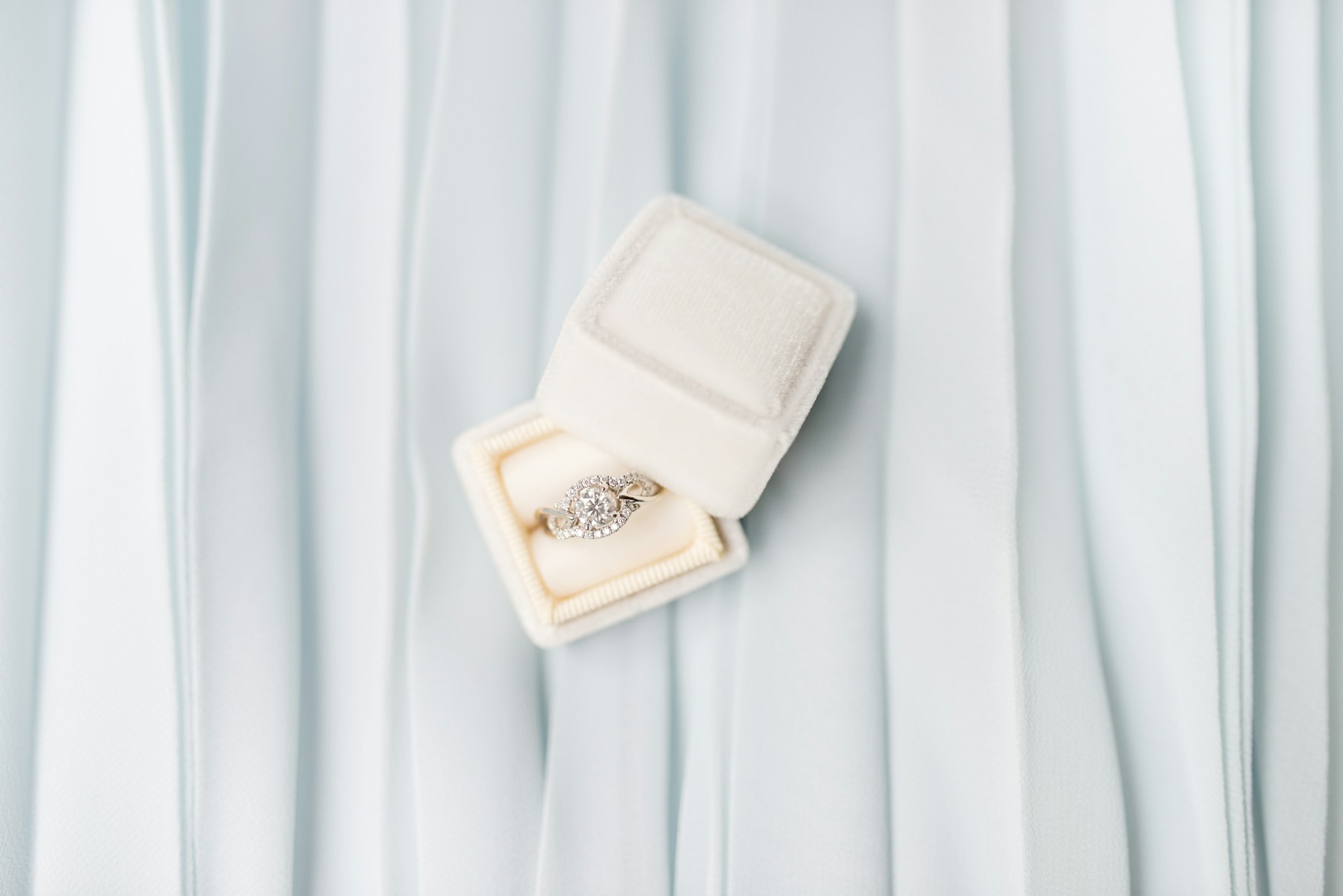 A white ring box holding an elaborate halo engagement ring on a pale blue fabric surface.