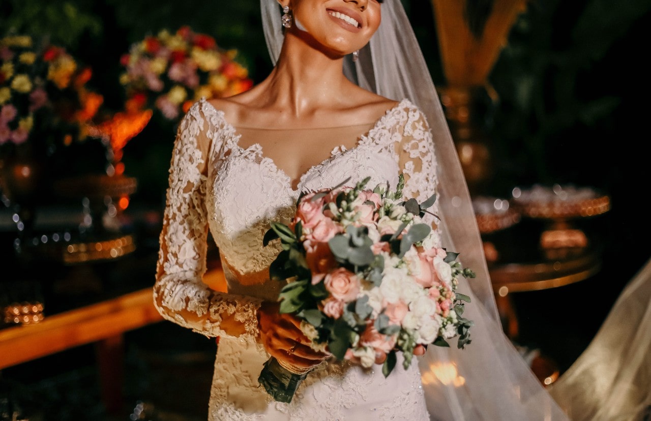 A bride making her entrance into her reception with a long sleeve, white lace dress, long veil swept over her updo hair and carrying a blush pink boutique of flowers