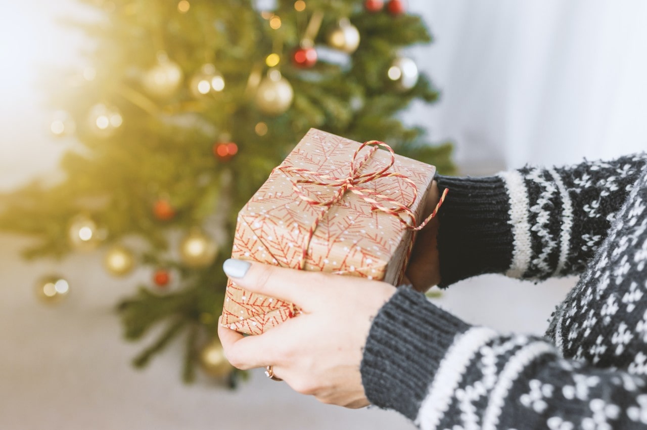 A woman holds a present of jewelry from Rottermond Jewelers wrapped just for her while she stands in front of a holiday tree while wearing a gray and white sweater.