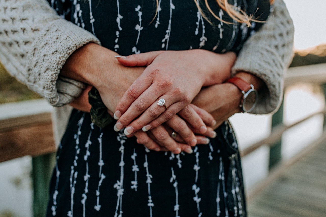 A close-up of a new bride-to-be wearing an engagement ring, embraced from behind on a bridge.