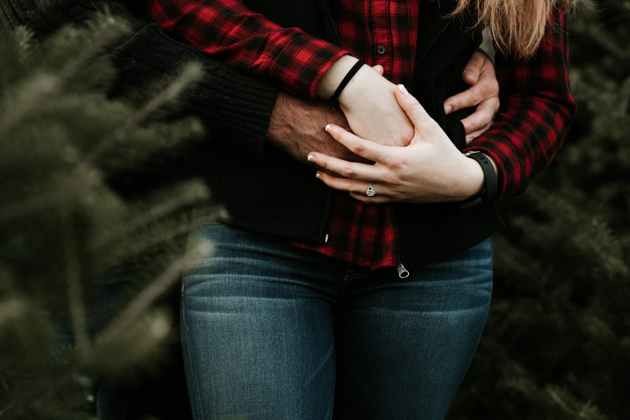 A close-up of a comfortably dressed couple embracing among pine trees, an engagement ring on her finger.