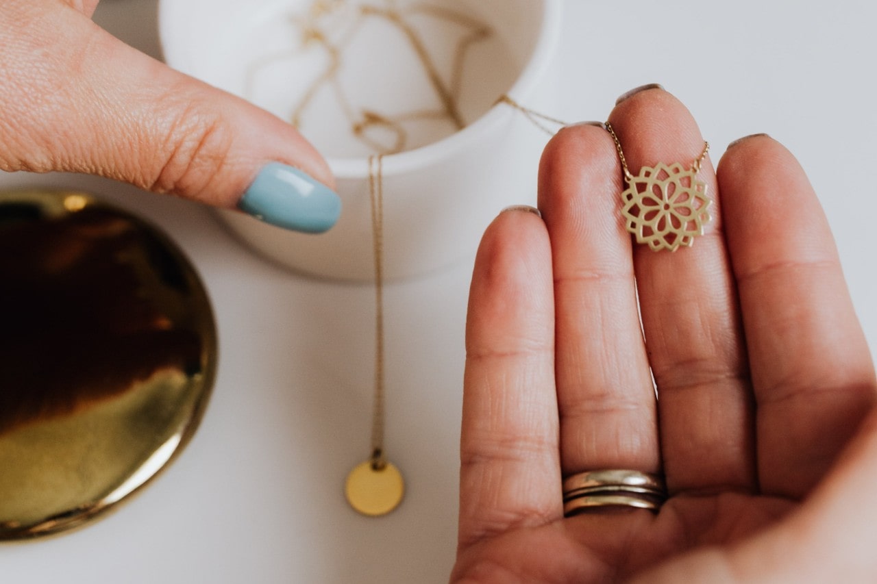 A close-up of a woman’s hands adorned with simple rings, selecting a dainty necklace from a small jewelry box.