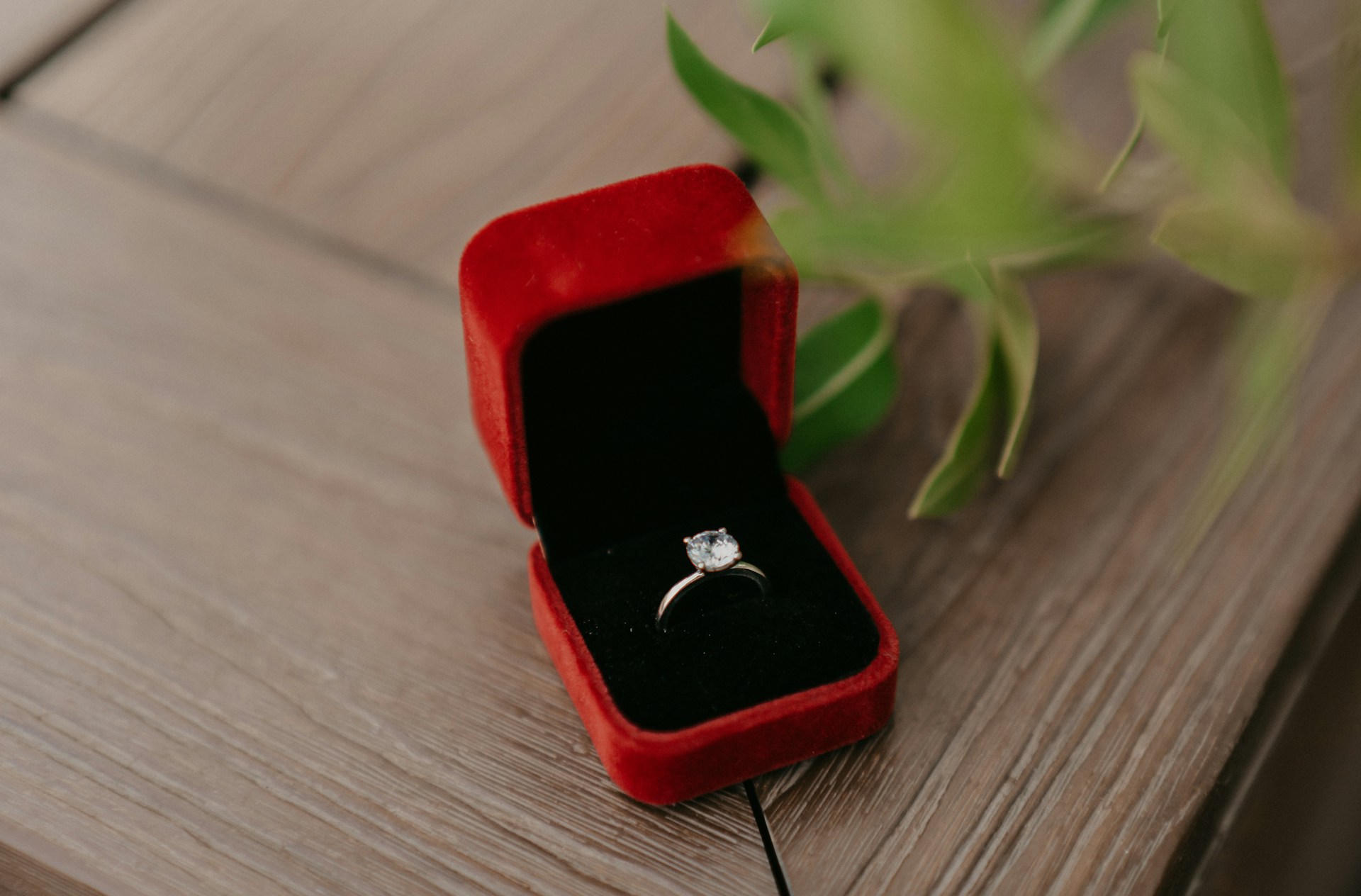 A close-up of a dainty solitaire engagement ring displayed in-box on a wooden tabletop.